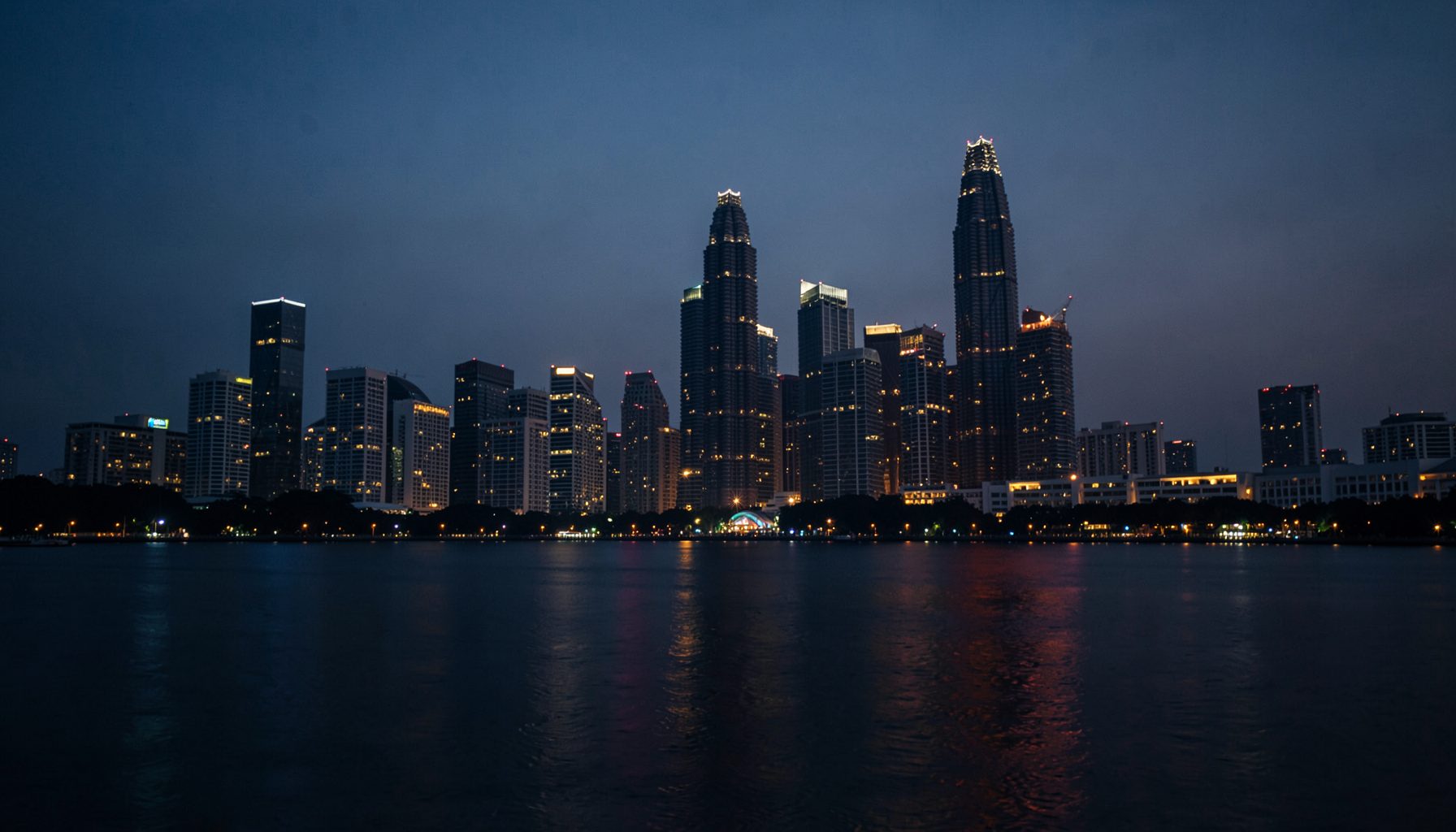 A dark, atmospheric view of Singapore's skyline at dusk with glowing city lights reflecting off calm water, conveying a mood of quiet reflection and financial contemplation.
